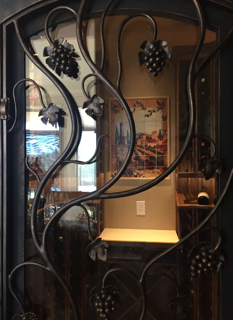 Glass wine cellar door with wrought-iron grapevine design, view of wine racks, bottle and tile vineyard mural inside