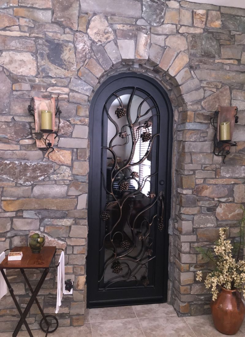 Arched wrought-iron door with grapevine motif set in rustic stacked stone interior wall, flanked by candle sconces