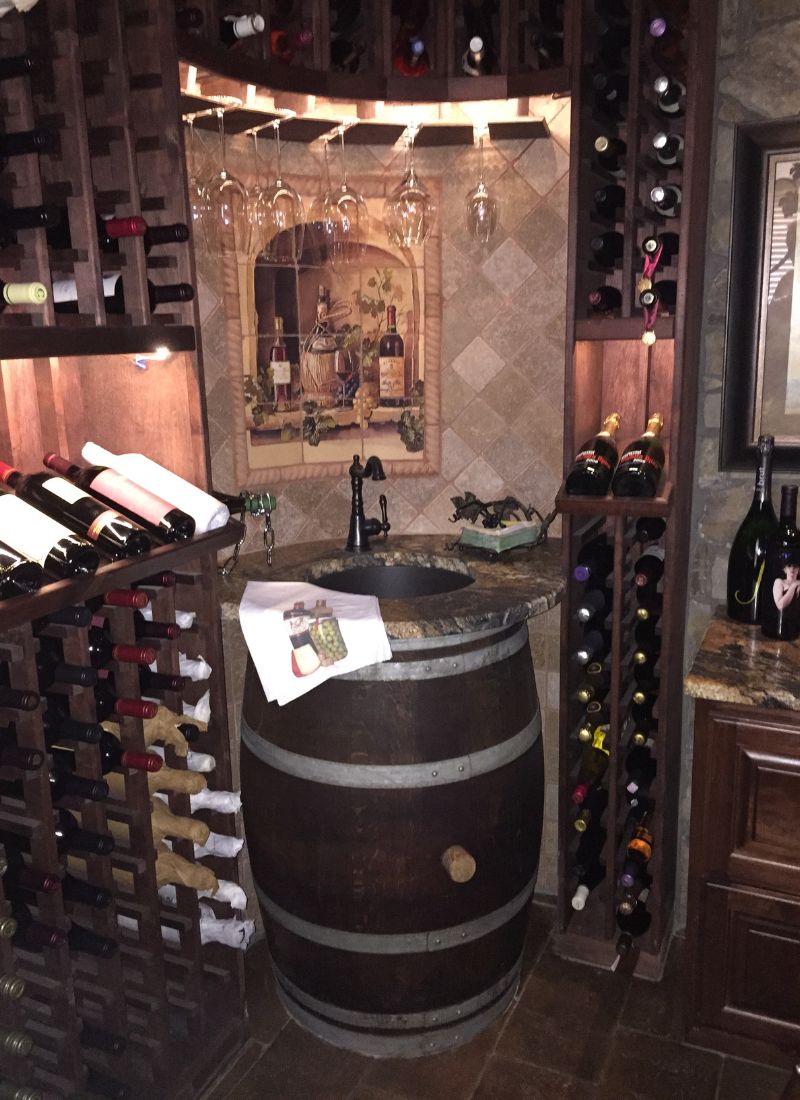 Wine cellar with wooden racks, wine bottles, hanging glasses and barrel sink beneath decorative tiled mural