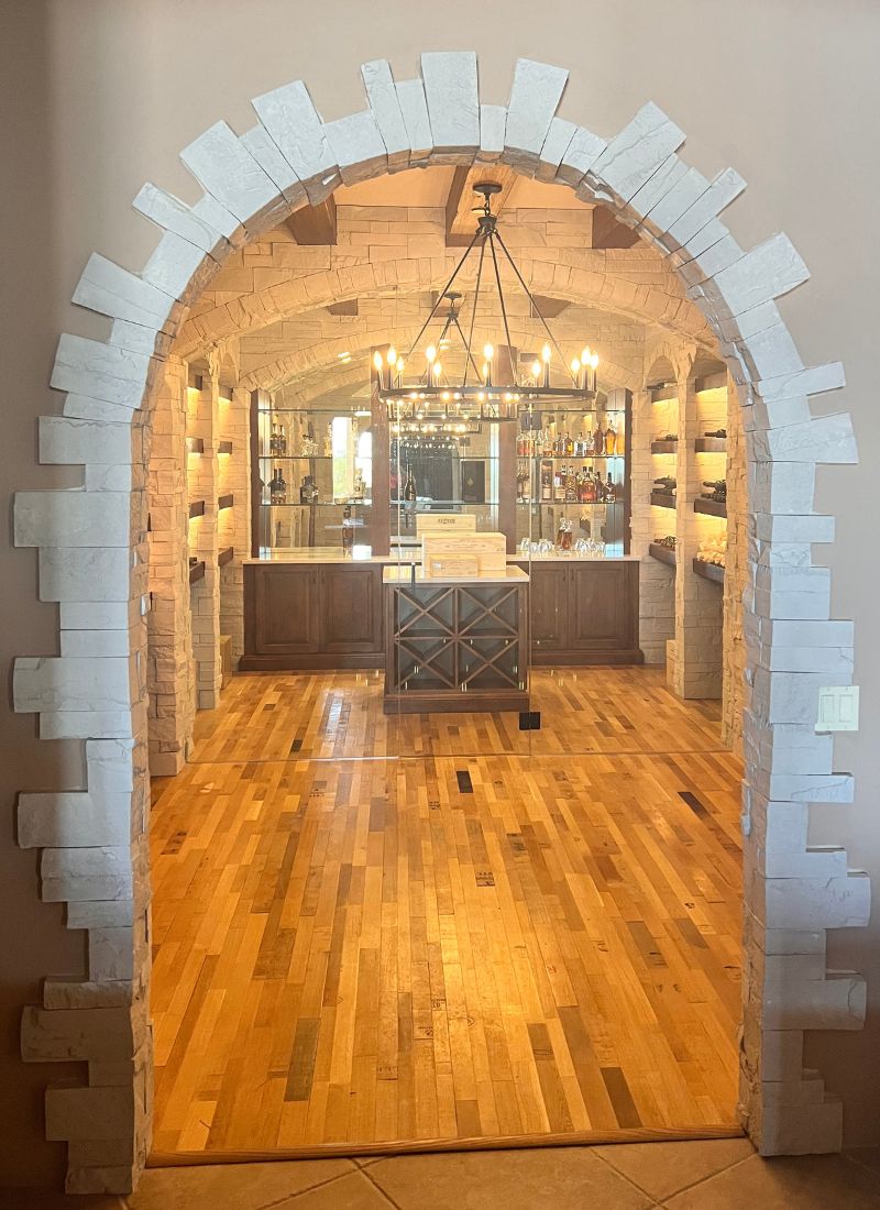 Stone arched entrance to elegant wine cellar tasting room with wooden floors, chandelier, wine racks and a central bar.