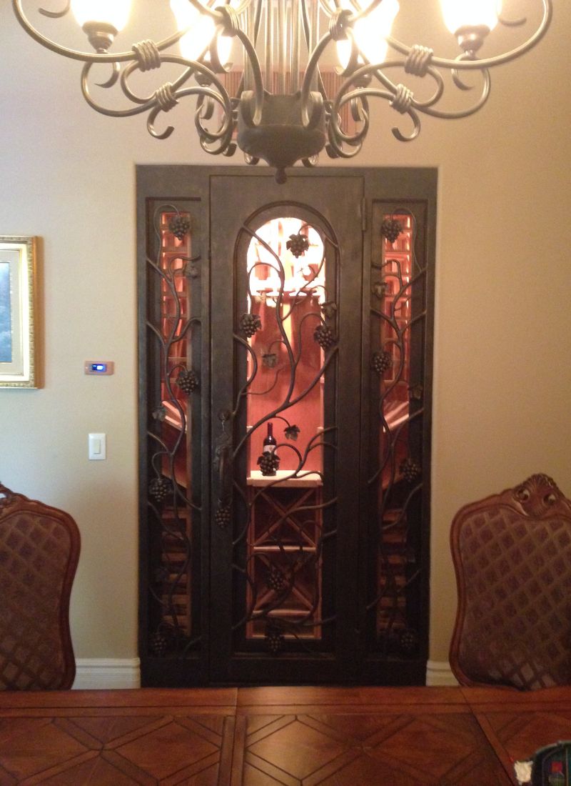 Illuminated wrought-iron grapevine wine cabinet with glass doors in elegant dining room under chandelier