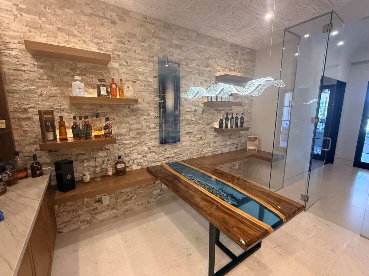 Modern home bar with epoxy resin river wood table, stone accent wall, floating shelves with liquor bottles and glass enclosure