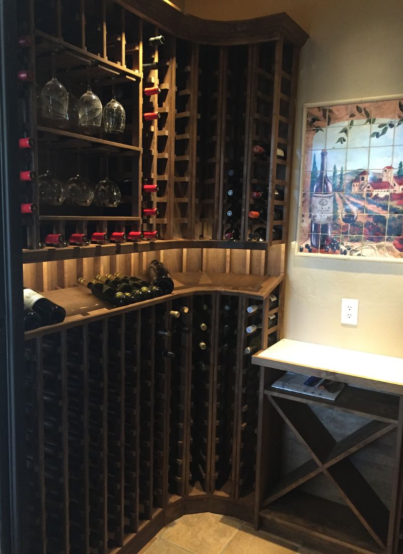 Corner wine cellar with tall wooden wine rack stocked with wine bottles, hanging glasses and a tiled wine mural.